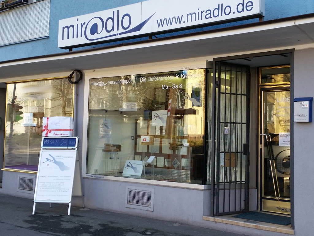 Vorm Schaufenster unser Plakatständer, auf dem oben ein Paket steht. miradlo-Versanddepot in Konstanz
