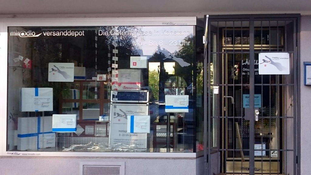 Schaufenster mit Ladentür, vor dieser ein Gitter, an der Tür ein Schild, im Fenster hängen Pakete