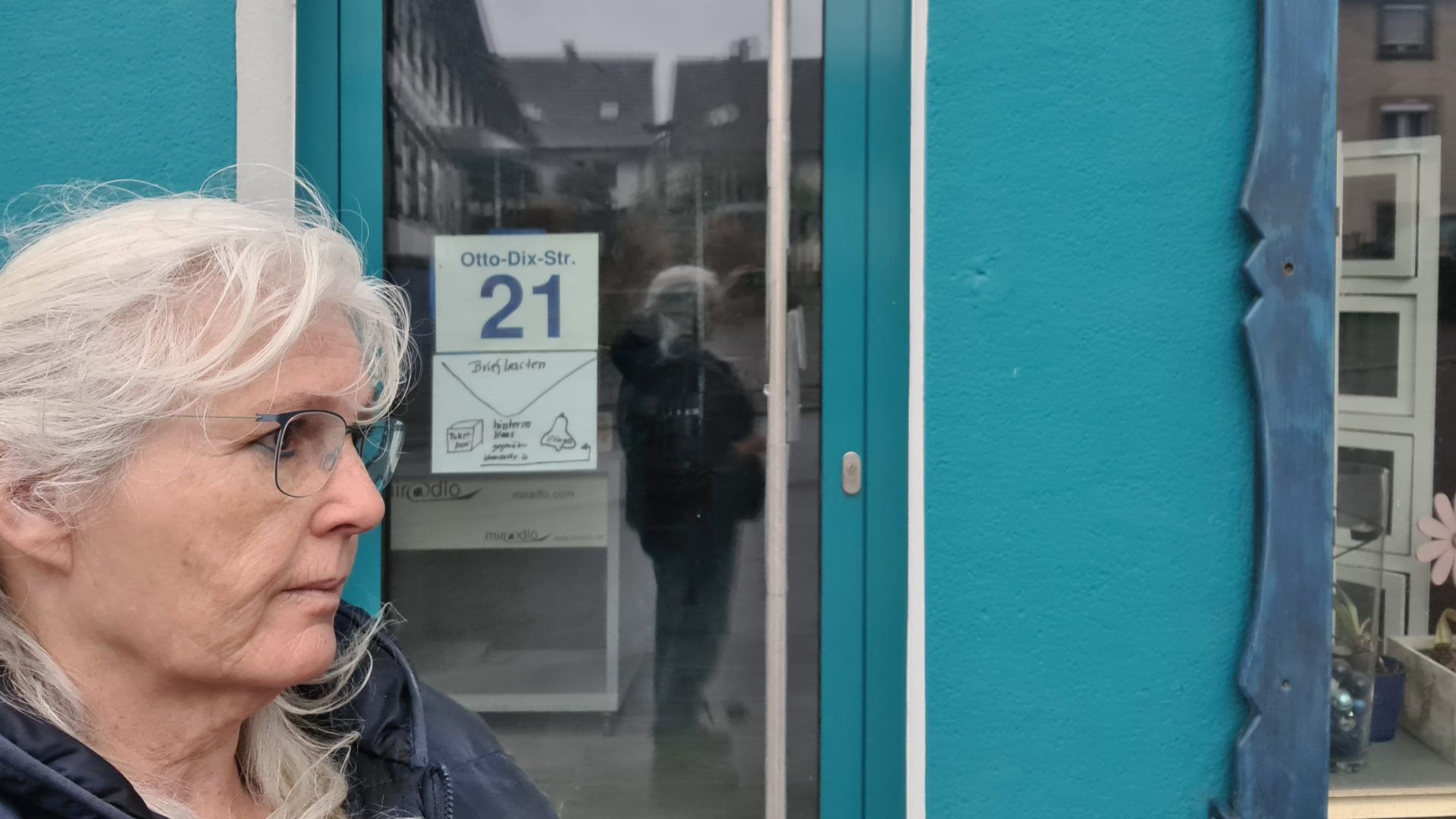 Frau mit weißen Locken, ziemlich sauer im Profil vor einer Glastür die Hinweise für Zusteller und Besucher zur Rückseite enthält