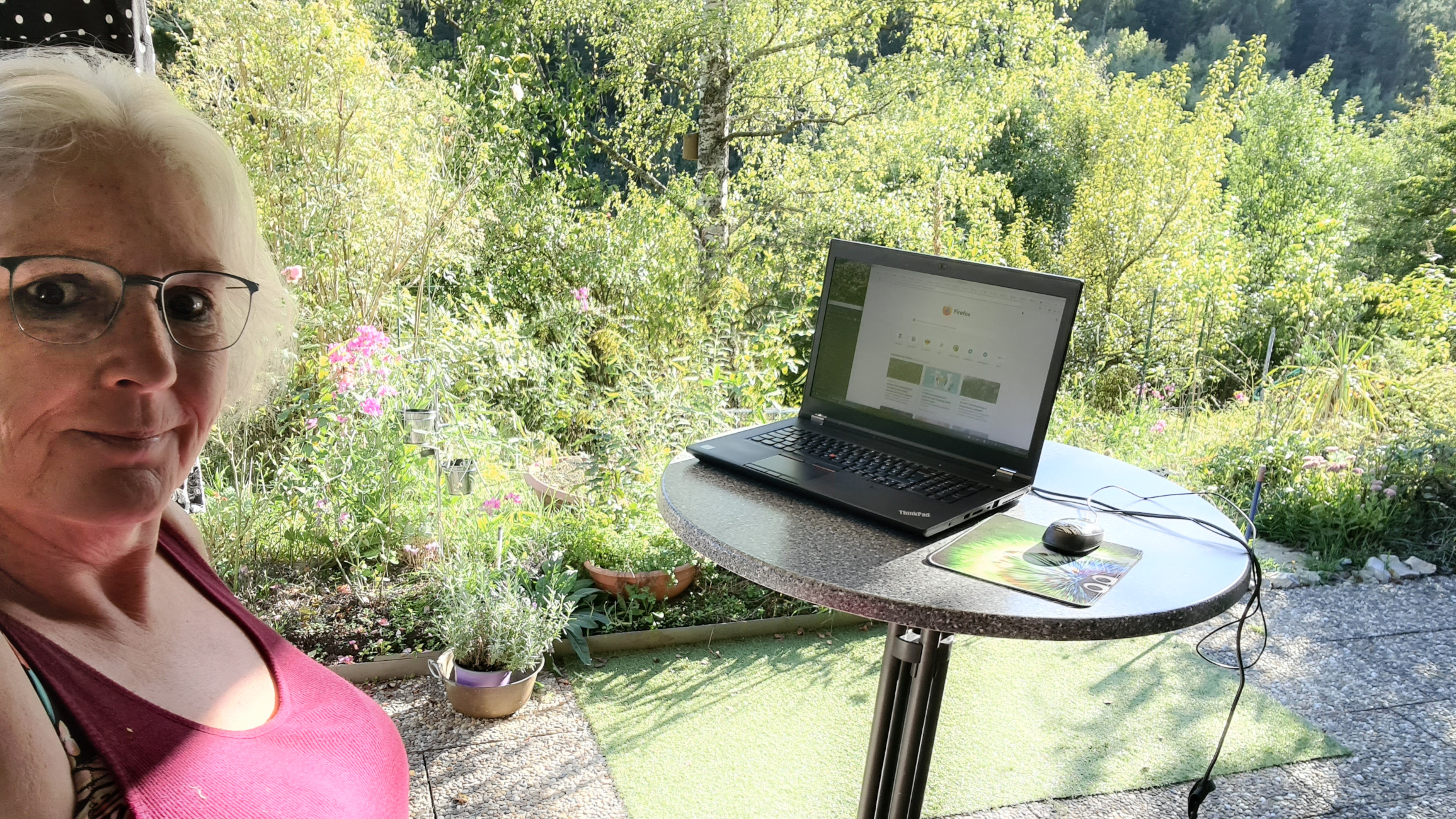 Frau mit langen weißen Locken lächelt auf der Terrrasse vor einem sehr grünen Garten, im Vordergrund ein Stehtisch mit großem Laptop