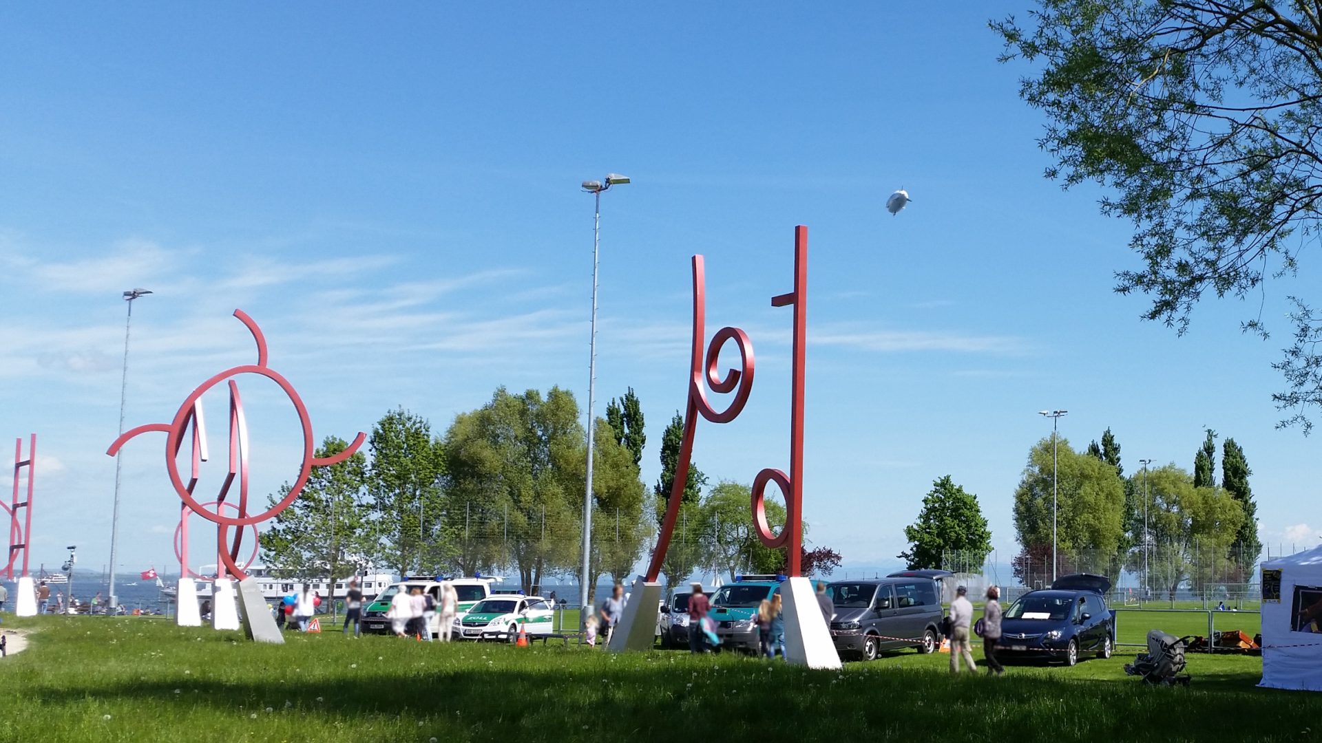 Messe, parkende Fahrzeuge, auch des deutschen Zolls am Rand einer grünen Wiese, vorn die verschiedenen roten Metallskulpturen der Kunstgrenze, im Hintergrund der See, ein Dampfer mit schweizer Fahne