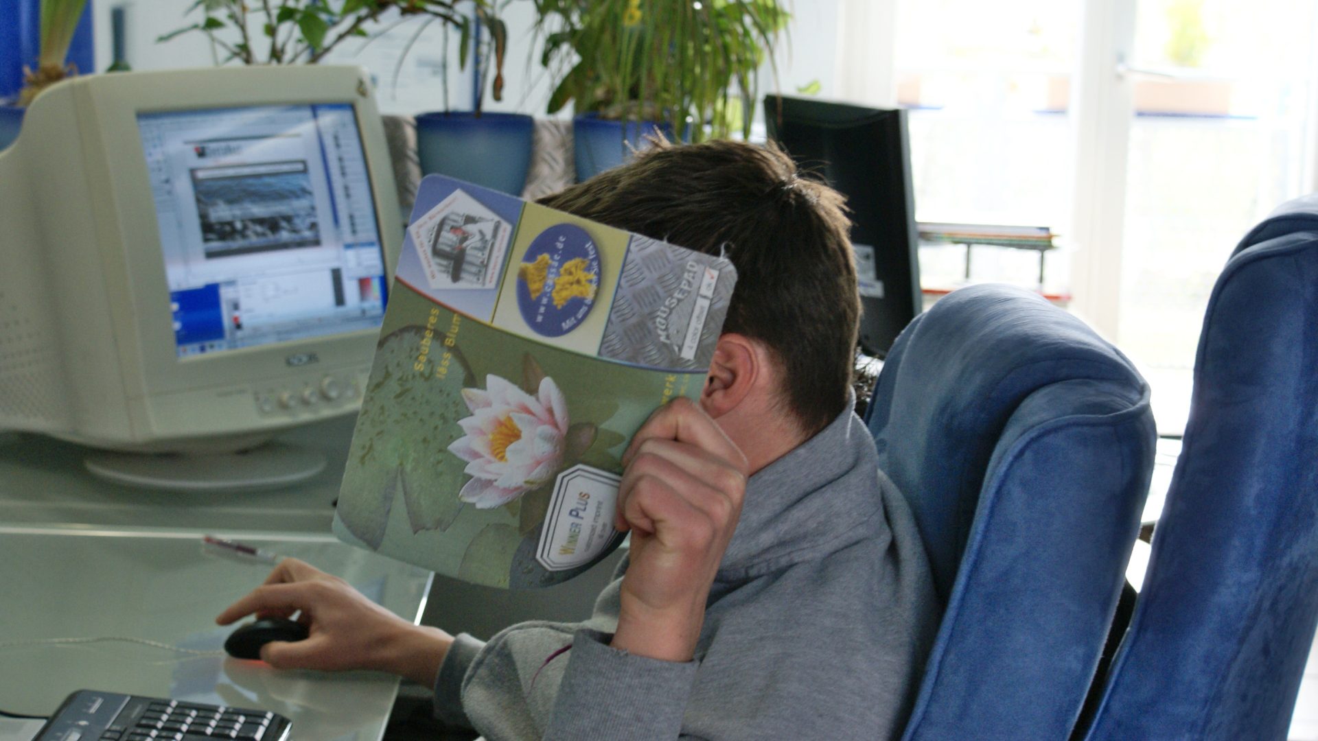 Junger Mann am Schreibtisch 2008 mit einem Riesenmonitor, hält sich ein Mauspad vors Gesicht und anonymisiert so das Bild perfekt..