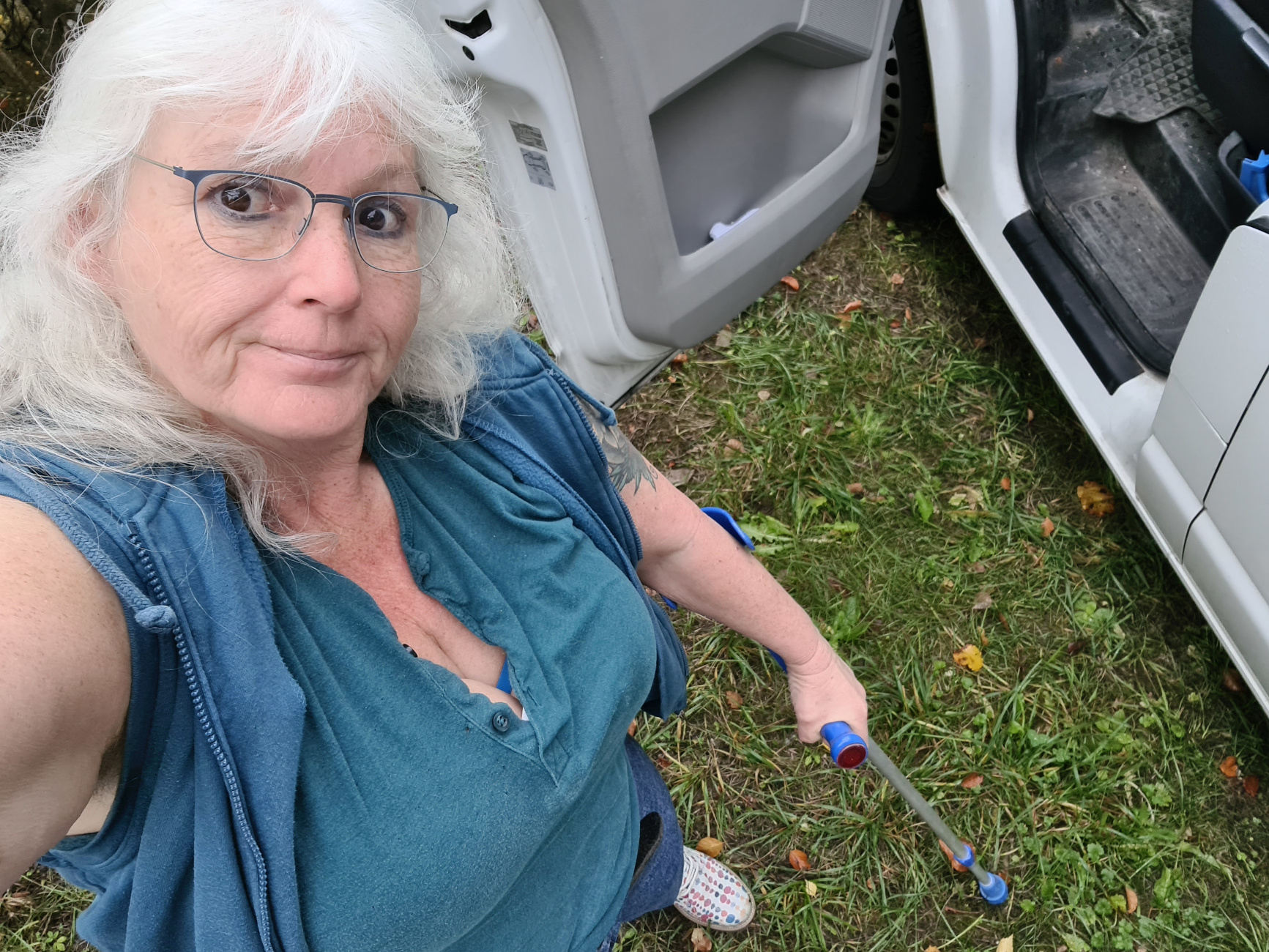 Frau mit langen weißen Locken und Brille schaut grinsend von unten in die Kamera, sie steht an einem Auto auf Wiesengrün und hält eine blau Krücke